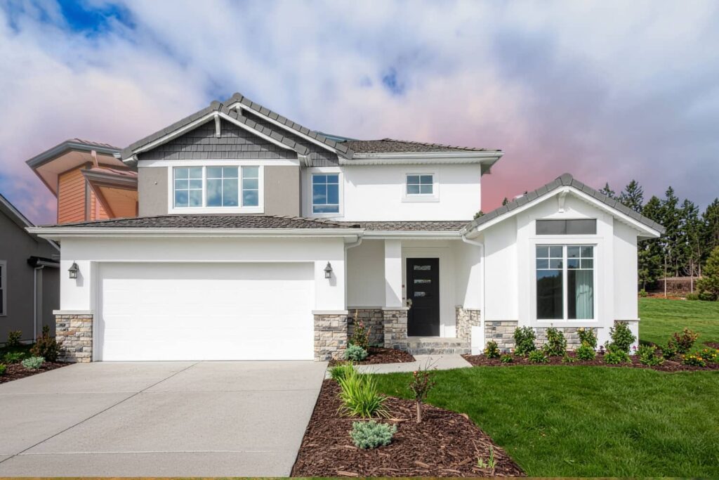 A modern, two-story modular home with white and grey siding, stone accents, and a two-car garage sits under a bright, cloud-filled sky.
